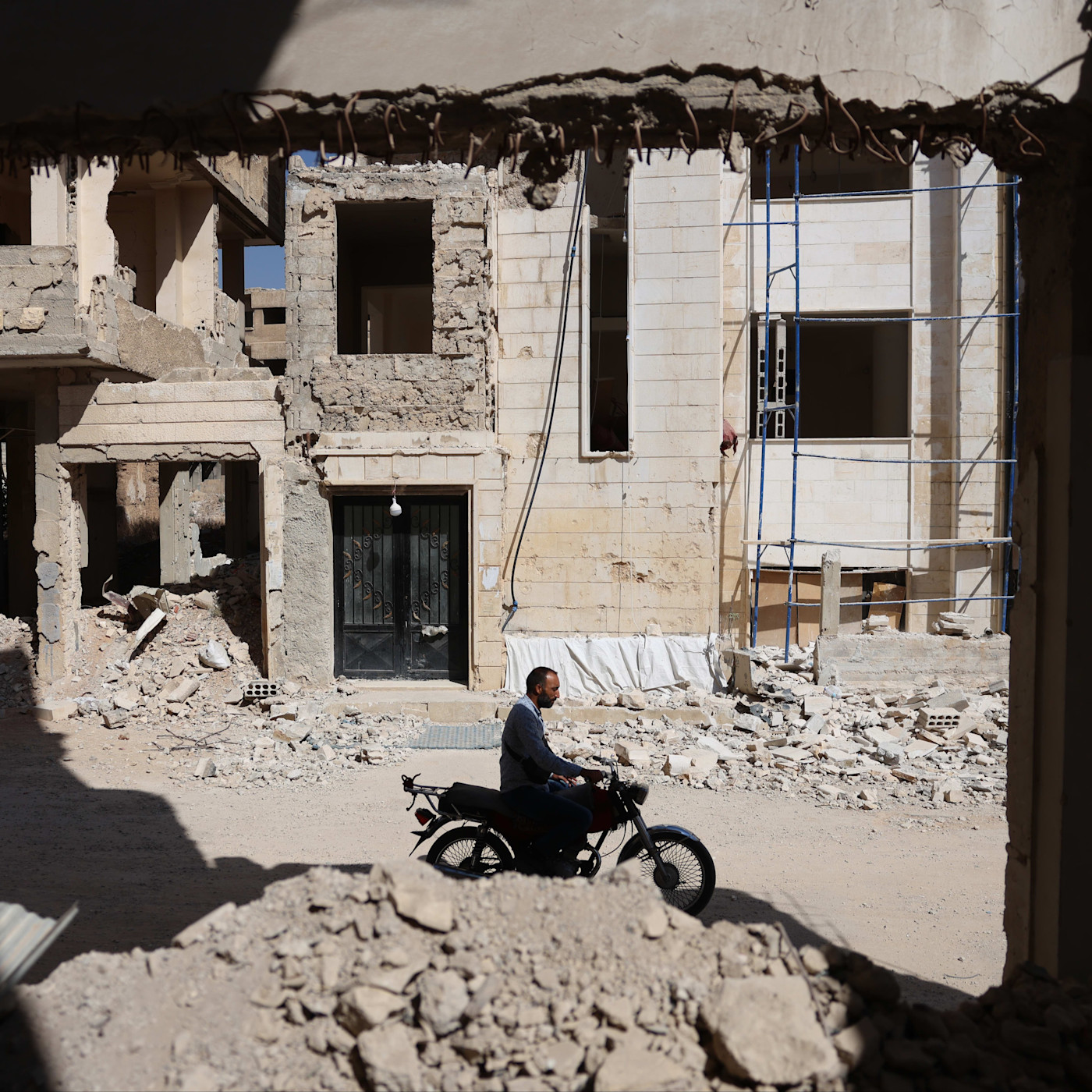 A man rides a motorcycle through a street filled with rubble, surrounded by heavily damaged and partially destroyed buildings.