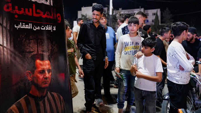 A group of people, including children, stand beside a poster showing Bashar al-Assad behind bars during celebrations in Idlib.