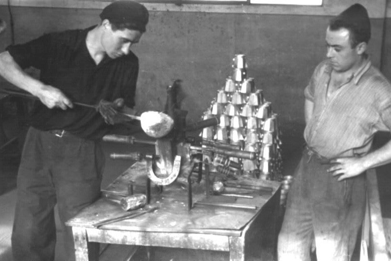 1950년대 두 작업자가 모카 익스프레스 커피포트를 만들기 위해 용융 알루미늄을 틀에 붓고 있다. 뒤에는 완성된 포트가 쌓여 있다.