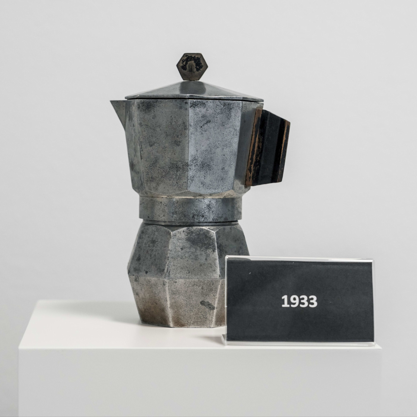 오래된 금속 마감의 빈티지 비아레티 모카 익스프레스 커피메이커가 "1933"이라고 적힌 표지판 옆 받침대 위에 전시되어 있다.