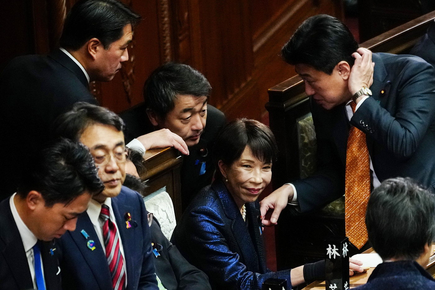 사나에 다카이치가 앉아 미소 지으며 일본 국회 총리 지명 투표 중 의원들에게 둘러싸여 있다.
