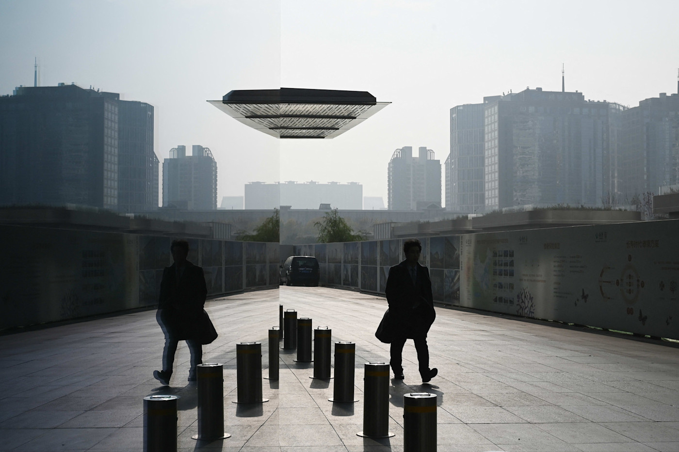코트를 입은 한 남자가 건물 밖 금속 볼라드 옆을 지나 걷고 있으며, 거울처럼 반사되는 표면에 그의 모습이 비쳐 보인다. 배경에는 고층 빌딩들이 보인다.